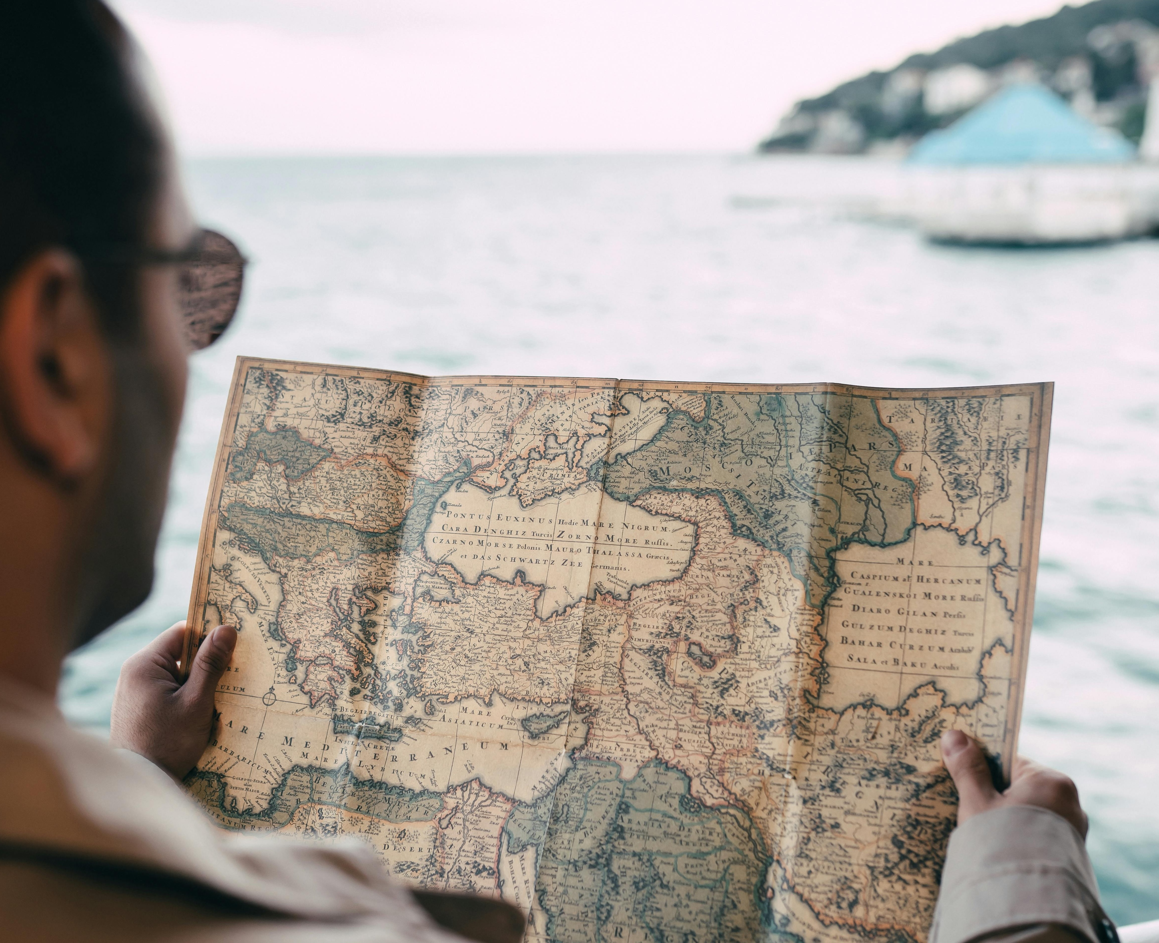 A person is holding an antique treasure map while looking out over a body of water. The map is detailed with various geographical features and marked paths, suggesting a journey to find hidden treasures. In the background, there is a blurred view of the shoreline and a building with a blue roof. The image evokes a sense of exploration and discovery.